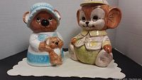 Photo of two ceramic cookie jars, a mother bear holding a small bear cub and a mouse in a hat and jacket, placed side by side on a white mat.