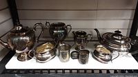 Full view of all silver plated items arranged on white shelf including two teapots, sugar bowl, creamer, urn, tumbler and two lidded containers.
