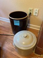 Photo showing two ceramic crocks: a large dark brown crock and a smaller light beige crock with lid, placed on hardwood floor near electrical outlets.