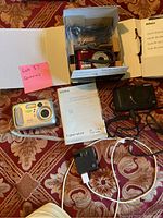 Three cameras displayed together: red Nikon Coolpix L24 in box with cables and manuals; silver Kodak EasyShare C310 with wrist strap; black Sony Cyber-shot with cables and manuals around them.
