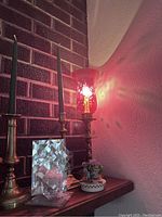 Candles and candle holders displayed on shelf with some lit; includes red glass hurricane candle holder, taper candles in brass holders, and clear geometric candle holder.