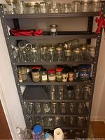 Gray metal shelving unit with multiple clear glass canning jars and spice jars, some jars lying on their side, some upright with metal lids.