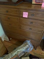 Front view of wooden chest of drawers showing six drawers and metal round knobs, wood in brown finish, placed on wood floor with some items nearby.