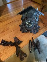 Photo of a dark wood carved cuckoo clock with two separate carved wooden leaves shaped pendulum pieces and two pine cone metal weights on the floor next to it.