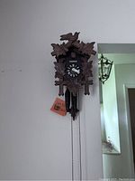 Front view of the wooden cuckoo clock mounted on the wall showing carved leaves, bird figure, black clock face with white numbers, and hanging chains with pinecone weights.