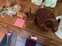 Photo showing a variety of decor items including a hand-painted ceramic cow, a small carved wooden animal, and a brown ceramic basket with handle on a wooden dresser.