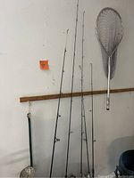 Four fishing poles and one fishing net arranged vertically against a white wall with a wooden rail.