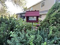Front view of wooden garden birdhouse with red roof on metal pole surrounded by greenery.