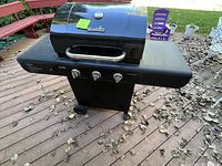 Black Char-Broil gas grill on outdoor deck with three control knobs, side shelves, and wheels.