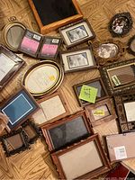 Various picture frames of different shapes and sizes arranged on a wooden floor, including wooden and ornate styles with some unopened frame mats.