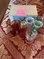 Photo showing various carnival glass items including amber sugar and creamer set with tray, colored ruffled edge cups, red glass pitcher, clear vase, and silver-top shaker, arranged on a patterned fabric.