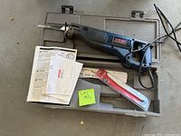 Ryobi Sawzall reciprocating saw with power cord placed on gray molded plastic carrying case. Includes instruction manuals and a sealed red blade package. The saw is visibly used, and the lot is marked with a sticky note 'LOT 125'.