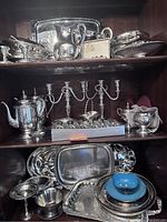 Full view of upper and middle shelves showing silver plated trays, teapot, candle holder, and various serving dishes in a display cabinet.