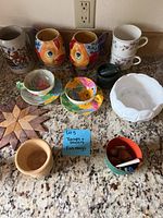 Photo showing assorted teacups, saucers, mugs, black mortar and pestle, wooden trivet, and a small red dish with clips and pens on a stone counter.