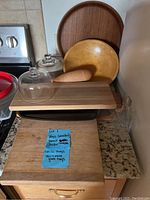 Photo showing wooden trays, small wooden cheese holder with pestle, wooden cutting board, a glass dome, and a small glass bottle on kitchen counter