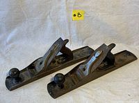 Two antique Stanley No.8 wood planes placed on light cloth background, showing side and top views, displaying metal bodies, wooden handles and knobs, with surface rust and wear.