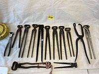 Top view showing the full lot of 14 antique blacksmith nippers arranged on a white cloth, varying sizes from small to very large
