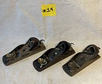 Three Stanley block planes displayed side by side on a fabric surface, showing metal bodies with black painted lever caps and adjustment knobs, all with visible wear and patina.