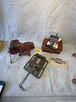 Lot of three vises arranged on light cloth backdrop: Craftsman 3.5-inch bench vise, Vacu Vise on wooden base, 4-inch drill press vise with adjustment screw.