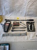 Overall view of the tools laid out on a white cloth background showing ratchet, wrenches, sockets, extension bar, socket set in packaging, and puller.