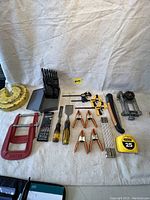 Photo showing assorted hand tools spread on white cloth, including drill bits in case, chisels, clamps, tape measure, and metal clamps.