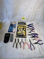 Full lot of assorted pliers, wire cutters, multitool with leather sheath, screwdriver bit set, and crimper tool taken on white backdrop.