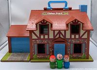 Front view of the Fisher-Price Little People Dollhouse showing Tudor-style exterior, blue garage door, roof handle, and three Little People figures in front.