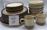 Stacked collection of plates and bowls along with 2 mugs showing the cream color with two brown rings near the base and dark brown rim edge.