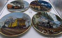 Four round collector plates featuring colorful steam train scenes in snowy and mountainous settings with gold rims.