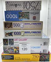 Stack of seven jigsaw puzzle boxes showing brands and piece counts.