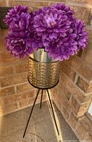 Front side view of the artificial purple flowers in the metal vase on the decorative stand against a brick wall.
