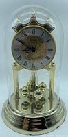 Full front view of Birks anniversary clock with glass dome showing brass base and columns, clock face with floral detail, and rotating pendulum balls underneath glass dome.