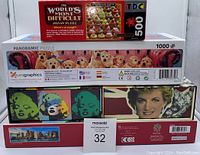 Stack of five puzzle boxes including The World's Most Difficult Cupcakes (500 pieces) on top, Eurographics panoramic yellow labrador puppies puzzle (1000 pieces) in middle, and three rectangular boxes featuring celebrity pop art and panoramic puzzles.