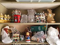 Photo shows a cardboard box containing various Christmas-themed decorations including multiple ceramic houses covered with snow effects, angel figurines, a red telephone booth ornament, a small figure of a girl in winter clothes, a Christmas-themed clock with reindeers, a wrapped glass jar or vase, a Christmas teapot, and wrapped knitted items.