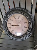 Front view of the antique round wall clock showing Roman numerals, black ornate hands, and decorative black frame with beaded edge.