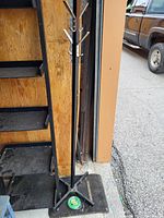 Full view of the black metal coat rack showing hook arms and four-legged base, placed on pavement next to wooden shelving unit and a vehicle.