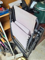 Folded folding chair with light beige fabric and black metal frame placed in a storage area next to gardening tools and pots.
