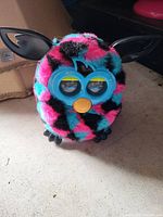 Colorful Furby toy with striped pink, blue, and black fur, blue face with yellow beak and ears, placed on carpeted surface.