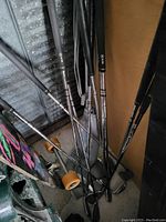 Photo of seven golf clubs with metal shafts leaning against a wall, showing a mixture of iron clubs and a putter with some visible brand markings on grips and shafts.