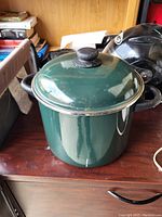 Photo of a large green metal stock pot with black handles and matching lid on a wooden surface, surrounded by various items in the background.