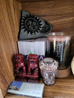 Photo of shelf with red Foo Dogs, decorative vase, candle, remote, box, and sun-face ornament
