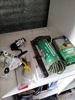 Photo of various outdoor items including fishing reels, an expandable garden hose, knives, and a camping kit on a white shelf.