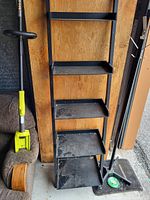 Front view of black metal shelving unit with five shelves showing dust and wear. Unit against wooden panel background with nearby objects such as a carpet cleaner and coat rack.