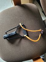 A handheld slingshot placed on a brown fabric chair seat, showing the black metal frame, black plastic handle, and yellow elastic bands with leather pouch.