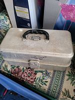 Closed gray molded plastic tackle box with black handle labeled 'Busy Boy Tackle Box' placed on floral patterned fabric.