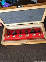Open wooden box showing six metal router bits in red holder with labels for bit types