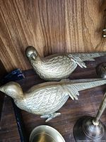 Photo showing two brass bird figurines with detailed feather engravings and long tails on a wooden surface.