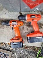 Two Black & Decker cordless drills, one with a 14.4V battery and the other with an 18V battery, laying on concrete next to a Canadian Tire plastic bin.