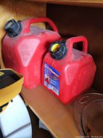 Photo shows two red plastic gas jugs on a wooden shelf. The larger jug is 10 liters and the smaller jug is approximately 5 liters. Both have black caps and handles for easy carrying.