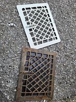 Two metal vent covers on pavement, one white and one rusted dark brown with lattice pattern and side latches.